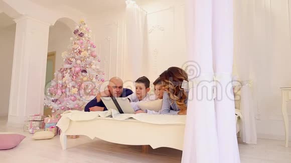 快乐的一家人快乐地在一起用电脑躺在明亮的卧室里有圣诞树和圣诞树视频的预览图