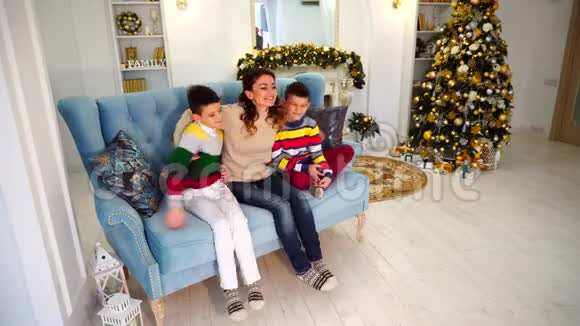 幸福的母亲两个儿子和男孩的兄弟并排坐在一起一家人抱在蓝色的沙发上装饰着节日视频的预览图