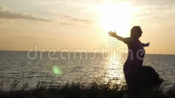 一个年轻的女孩正在草地上在夕阳和大海的背景下旋转享受着阳光4k3840x2160视频的预览图