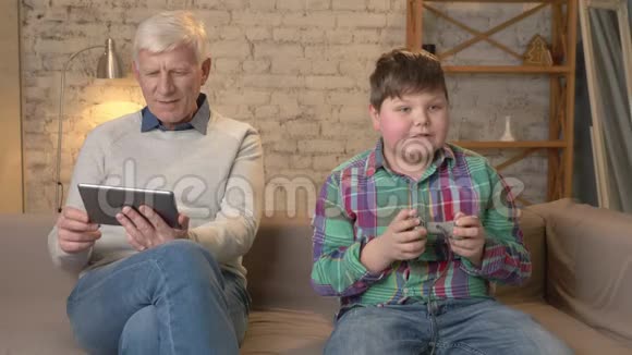 祖父和孙子坐在沙发上一个老人用平板电脑一个年轻的胖子在游戏机上玩视频的预览图