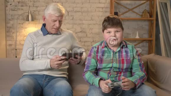 祖父和孙子坐在沙发上一个老人用平板电脑一个年轻的胖子在游戏机上玩视频的预览图