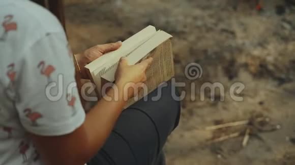 一个女孩坐在森林里看书女孩坐在火旁边的一根木头上读着一本书特写慢动作视频的预览图