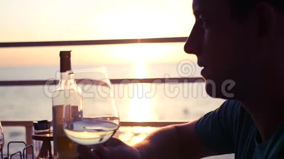年轻有魅力的男人正在一家咖啡馆里在大海和日落的背景下喝着一杯葡萄酒高清高清视频的预览图