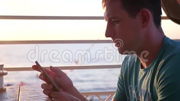一个年轻有魅力的男人坐在海边的日落时分手里拿着一个电话正用手指穿过视频的预览图