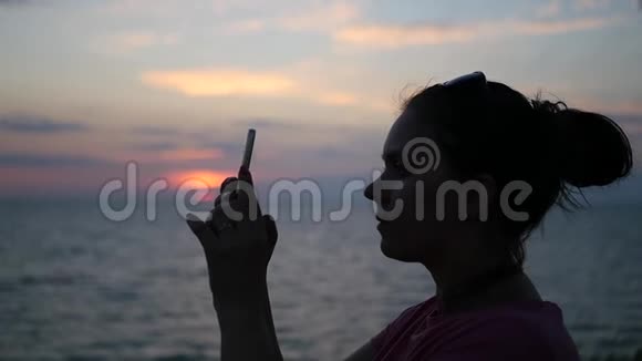 女孩在夕阳背景下站在海边的手机上拍照动作缓慢高清1920x1080视频的预览图