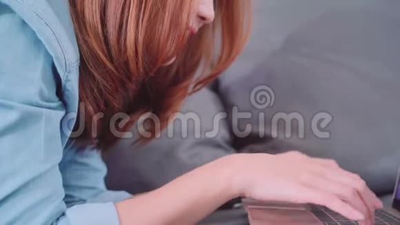一幅美丽迷人的年轻微笑的亚洲女人的肖像她在放松时躺在沙发上用电脑或笔记本电脑视频的预览图