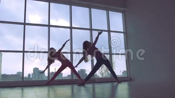 两位身材苗条的瑜伽女性在工作室里用自然光线练习伸展三角形姿势视频的预览图