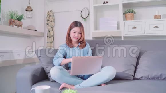 美丽迷人的年轻微笑的亚洲妇女使用电脑或笔记本电脑当躺在沙发上放松在客厅在家里视频的预览图