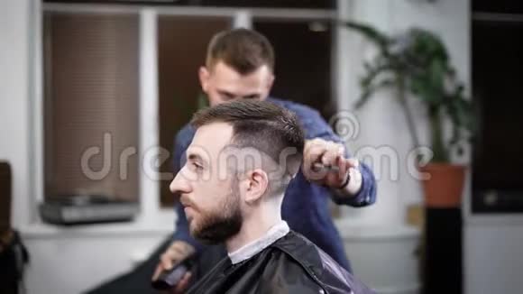 年轻的理发师站着在理发店里用剪刀给有吸引力的男人理发有胡子的人坐在上面视频的预览图