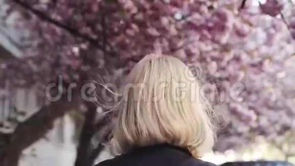 穿着皮夹克的漂亮金发女孩在城市里欣赏樱花转向相机迷人地微笑着视频的预览图