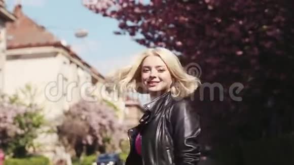 穿着皮夹克的漂亮金发女孩在城市里欣赏樱花转向镜头迷人地微笑着视频的预览图