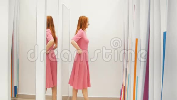 带眼镜的年轻女子试着在试衣间镜子旁穿着粉红色的裙子购物概念视频的预览图