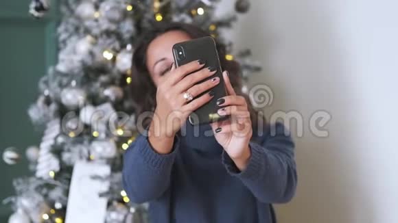 在圣诞树的背景下用手机拍下迷人的深色头发的自拍视频的预览图
