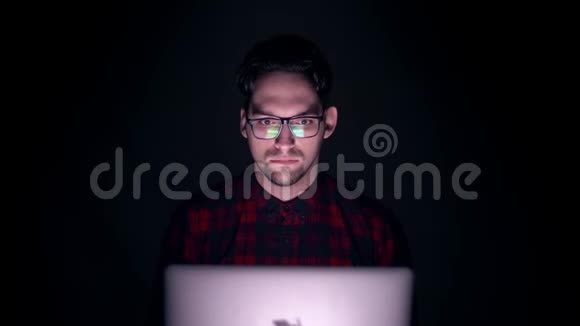 害怕的男人看起来像恐怖电影黑暗中监视器后面一个人的肖像一个男人在黑暗中看着电脑上的夜晚视频的预览图