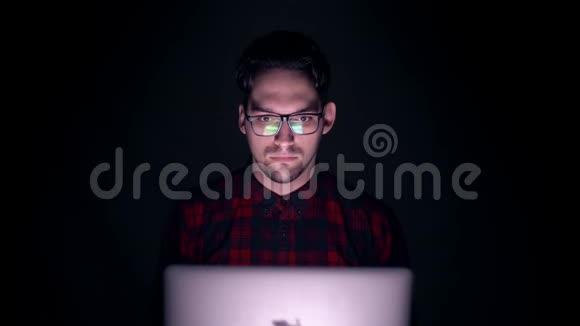 害怕的男人看起来像恐怖电影黑暗中监视器后面一个人的肖像一个男人在黑暗中看着电脑上的夜晚视频的预览图