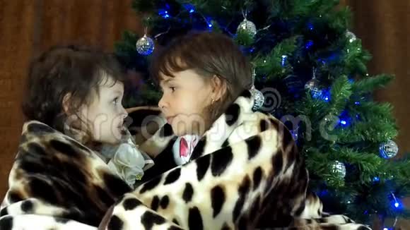 孩子们坐在圣诞树附近的地毯下两个小女孩被一条斑点毯子覆盖着两个姐妹在Ch下玩视频的预览图