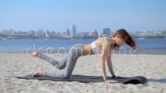 年轻苗条的运动女性在沙滩上做瑜伽练习和伸展运动以城市为背景健康生活方式视频的预览图