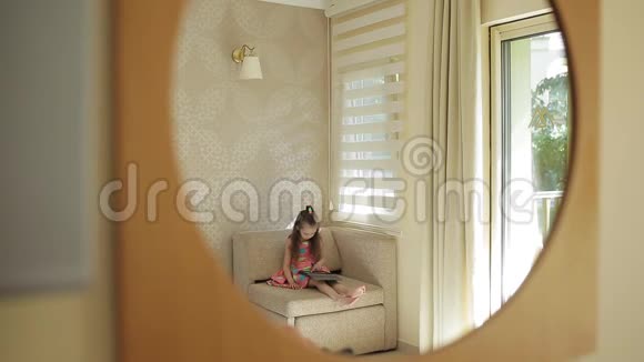 一个穿着漂亮明亮连衣裙的小女孩坐在窗边的房间里用平板电脑对着镜子看那个女孩视频的预览图