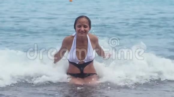 穿着漂亮比基尼的漂亮女孩坐在水上在鹅卵石海滩上举手泼水慢动作视频的预览图