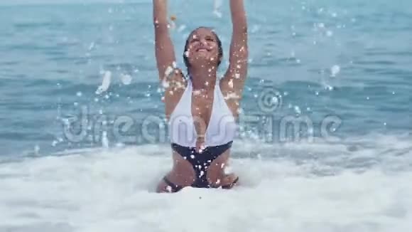 穿着漂亮比基尼的快乐年轻女子坐在海边的水里举起手来泼水在阳光明媚的日子里微笑视频的预览图