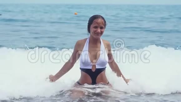美丽的女人穿着漂亮的比基尼坐在海边的水上举起手泼水在阳光明媚的日子里微笑慢慢地视频的预览图