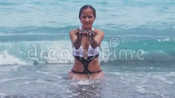 年轻可爱的女士穿着漂亮的比基尼坐在水中岸边洒满双手溅起水花视频的预览图