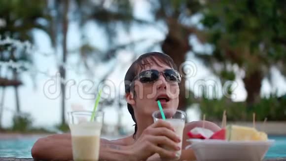 年轻的帅哥戴着太阳镜喝着鸡尾酒在游泳池里吃着热带水果旅行度假视频的预览图