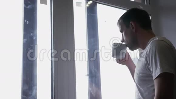 年轻迷人的男子穿着白色t恤喝着咖啡站在窗边望向远方4K3840x2160高清高清视频的预览图