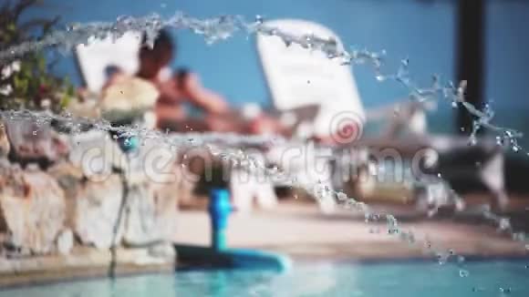 戴墨镜的年轻人躺在游泳池的日光浴床上前景上喷洒着水视频的预览图