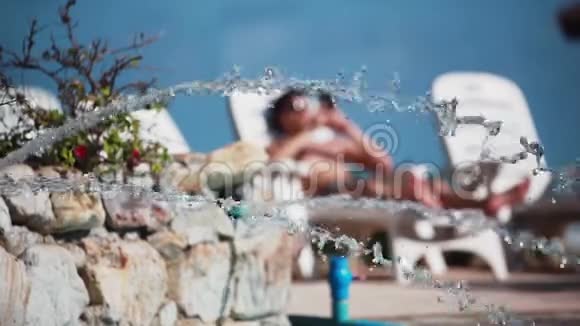 年轻人戴着太阳镜躺在游泳池的日光浴床上用手机喷水视频的预览图