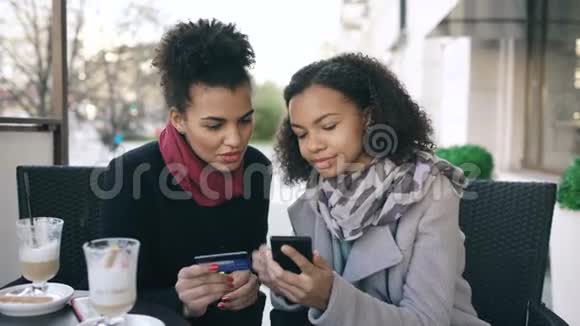 两位有魅力的混血女人一边聊天一边喝咖啡一边用信用卡和智能手机进行网上购物视频的预览图