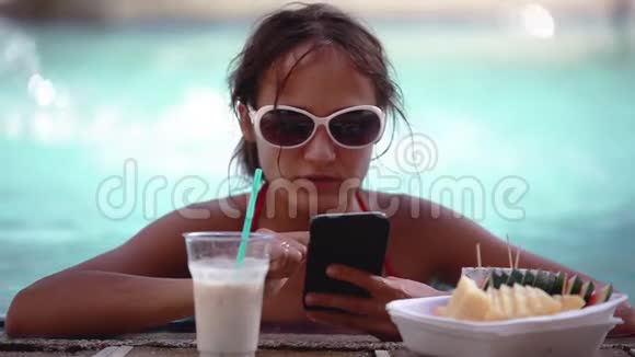 戴着太阳镜的年轻漂亮女人在游泳池里和手机饮料鸡尾酒聊天高清1920x1080视频的预览图