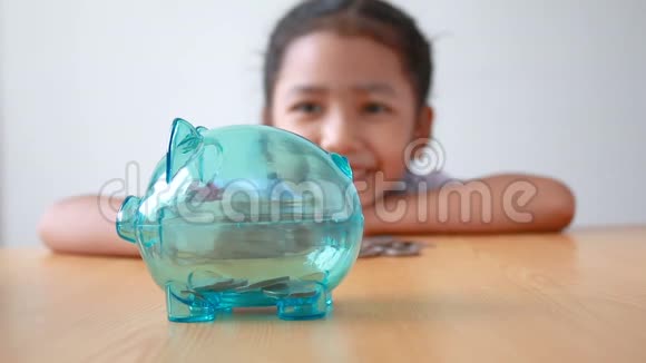 泰国幼儿园的亚洲小女孩穿着学生制服把钱币放在木桌上的透明存钱罐里比喻金钱萨夫视频的预览图