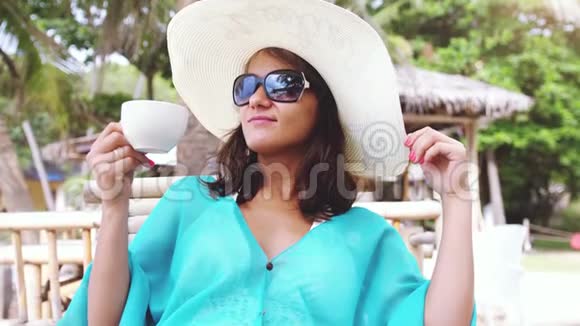 年轻优雅的黑发女人带着帽子和太阳镜度假坐在海滩咖啡馆欣赏海景视频的预览图
