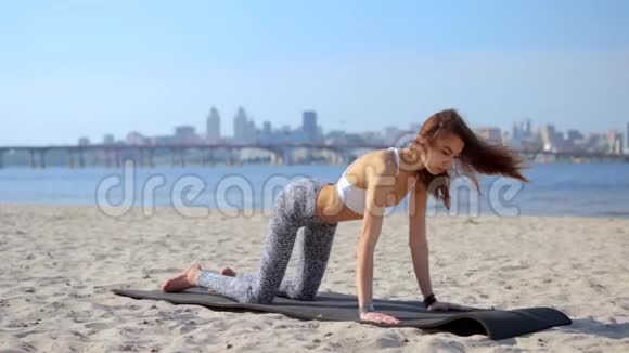年轻苗条的运动女性在沙滩上做瑜伽练习和伸展运动以城市为背景健康生活方式视频的预览图