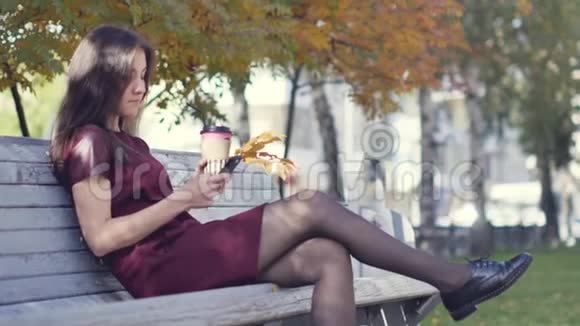 一个美丽快乐的年轻女孩外表迷人手里拿着手机坐在城市公园的长凳上视频的预览图