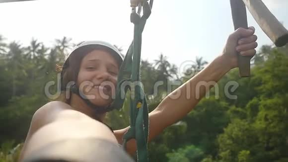 在美丽的热带丛林和阳光明媚的灯光下戴着头盔的年轻快乐的漂亮女人在高速的高空滑索视频的预览图