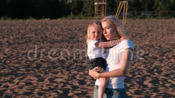 美丽的金发妈妈和女儿在日落时在海滩上拥抱和说话视频的预览图