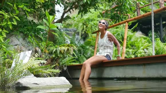 可爱快乐的年轻女人戴着太阳镜在瀑布旁的天然池塘里有鱼水疗慢动作1920x1080高清视频的预览图