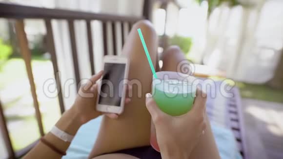 一位身穿泳装的年轻女子正在度假她躺在躺椅上手里拿着一杯鸡尾酒和一台手机视频的预览图
