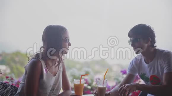 年轻漂亮的夫妇戴着太阳镜戴着智能手机坐在户外咖啡馆里聊天喝着鸡尾酒视频的预览图