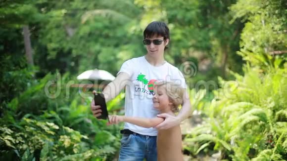 父亲戴着太阳镜和儿子在瀑布前用相机拍照泰国苏梅岛视频的预览图