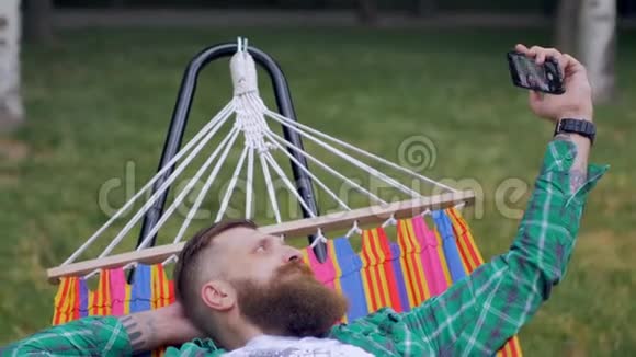 一个长着胡子的野蛮男人躺在大自然的吊床上在电话里自拍视频的预览图
