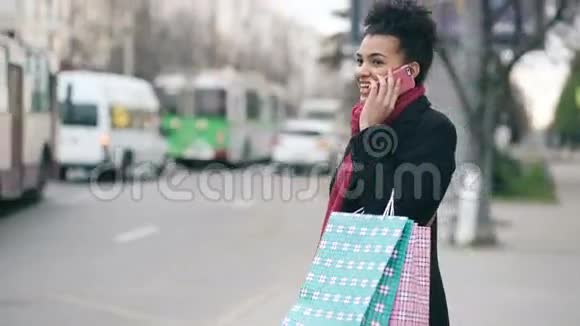 漂亮的非裔美国女人带着购物袋一边等出租车一边从商场回来一边打电话视频的预览图