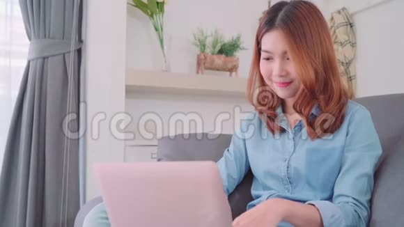 美丽迷人的年轻微笑的亚洲妇女使用电脑或笔记本电脑当躺在沙发上放松在客厅在家里视频的预览图