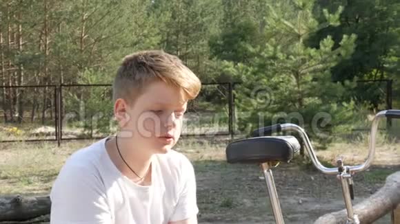 可爱的金发碧眼一个十几岁的男孩坐在自行车旁边的公园里在夏天说话情感肖像画视频的预览图