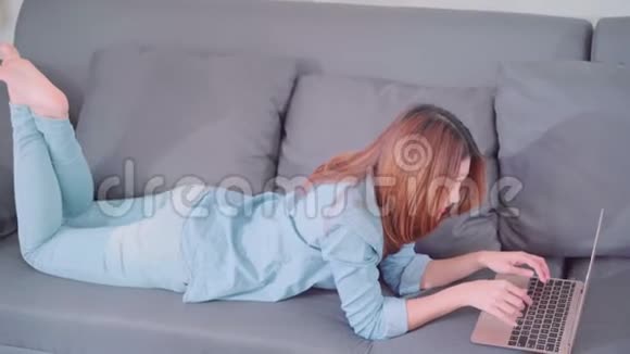 年轻微笑的亚洲女人在家里客厅放松时一边躺在沙发上一边使用电脑或笔记本电脑视频的预览图