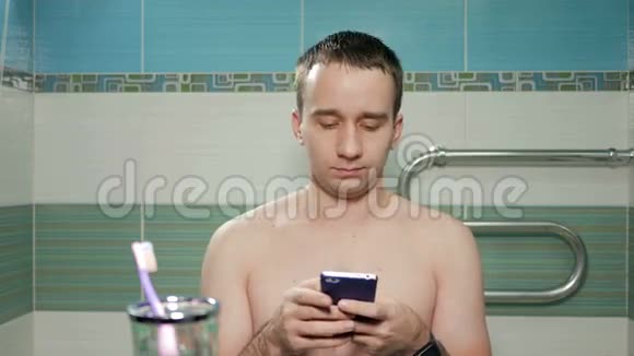 年轻迷人的男人在酒店房间的浴室里用电话写短信不穿有裸露躯干的T恤衫视频的预览图