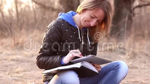 美丽的少女坐在公园秋天落叶上读着平板电脑在笔记本上做笔记视频的预览图