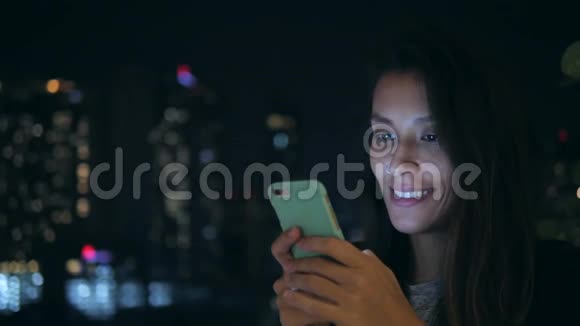 年轻的微笑女人在城市的夜晚在手机上发短信4K使用智能手机的混血女孩视频的预览图